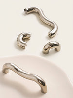 Nickel chrome arch knobs and wavy pulls scattered on cream tray, multiple angles