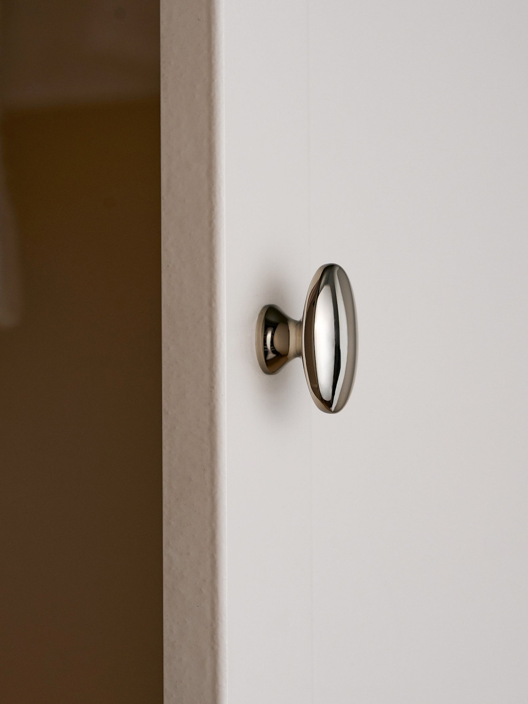 Nickel silver oval cabinet knob mounted on white cabinet door, edge view
