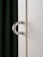 Polished chrome C-shaped cup pull on white cabinet door, green curtain background