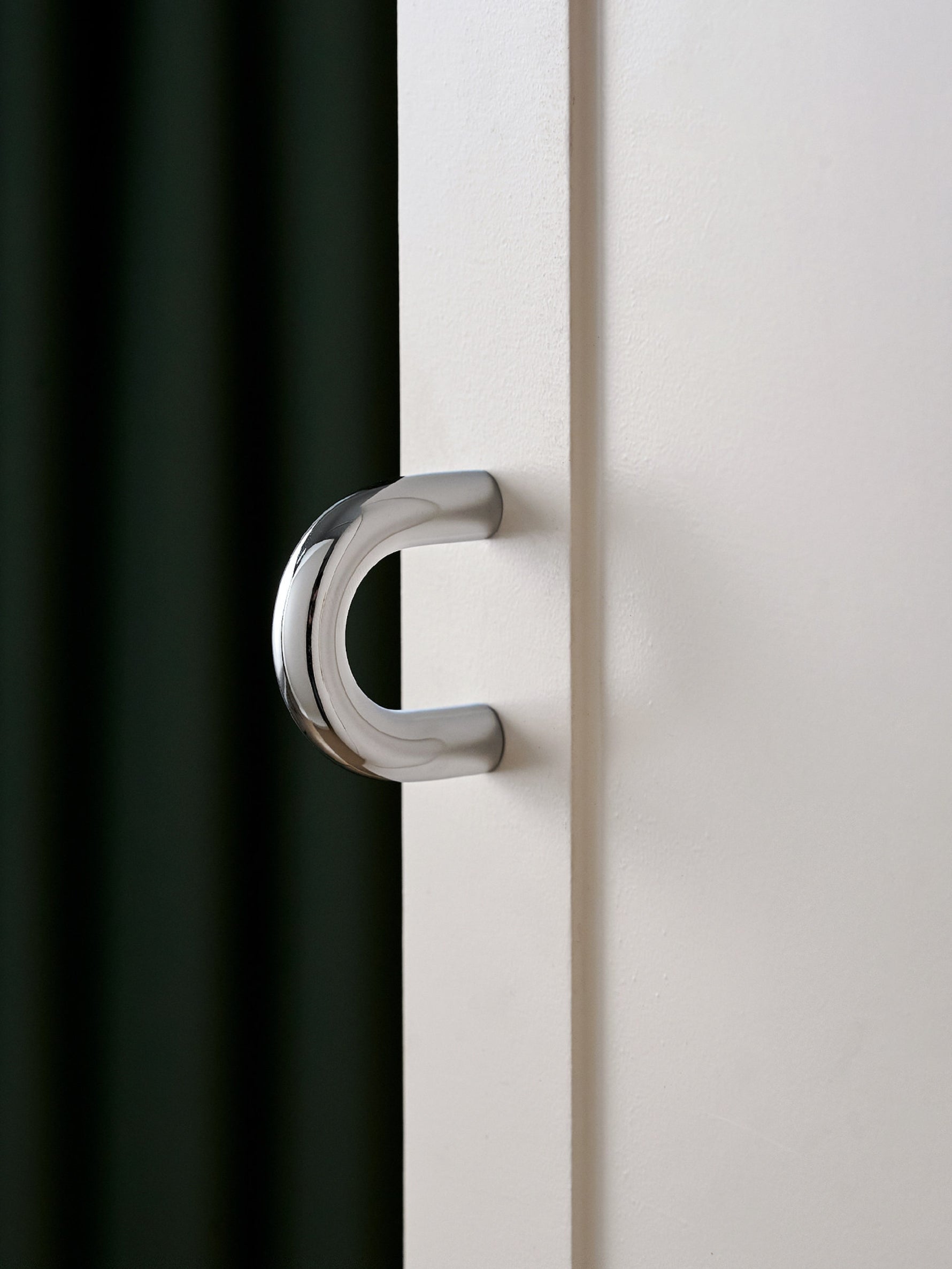 Polished chrome C-shaped cup pull on white cabinet door, green curtain background