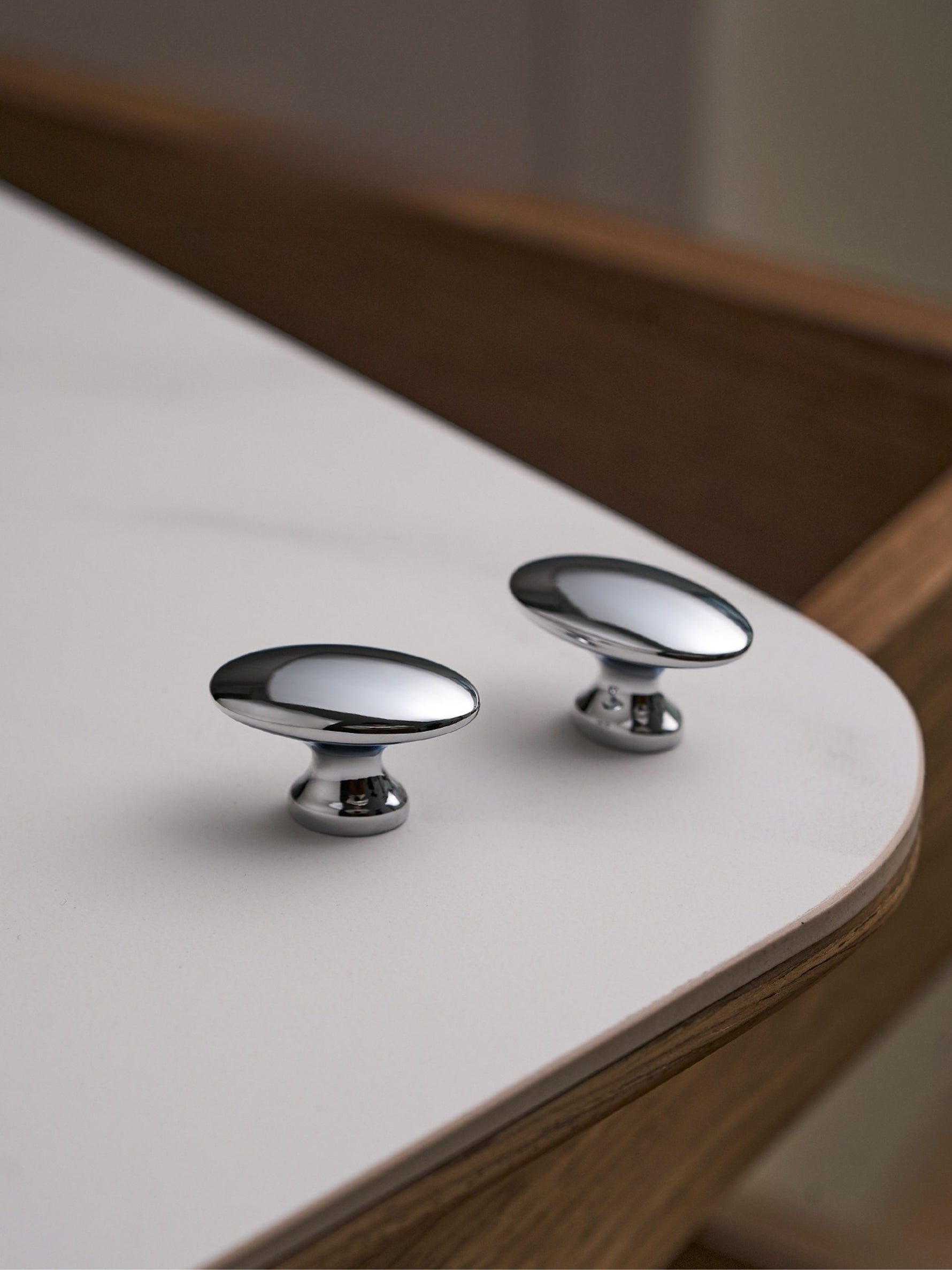 Two polished chrome oval knobs on white table with walnut edge