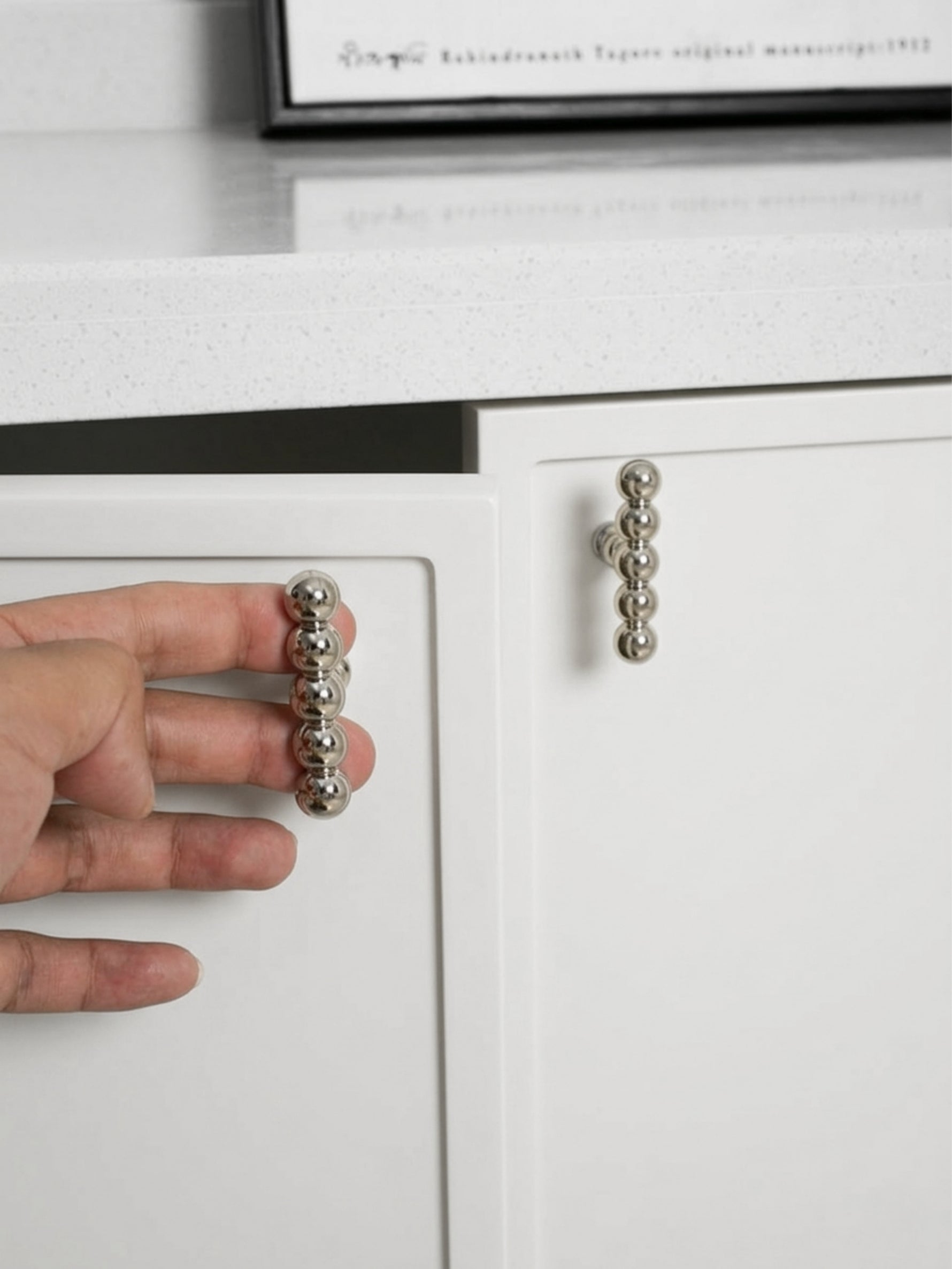 Hand holding a polished chrome beaded ball knob beside a second knob mounted on white cabinet door