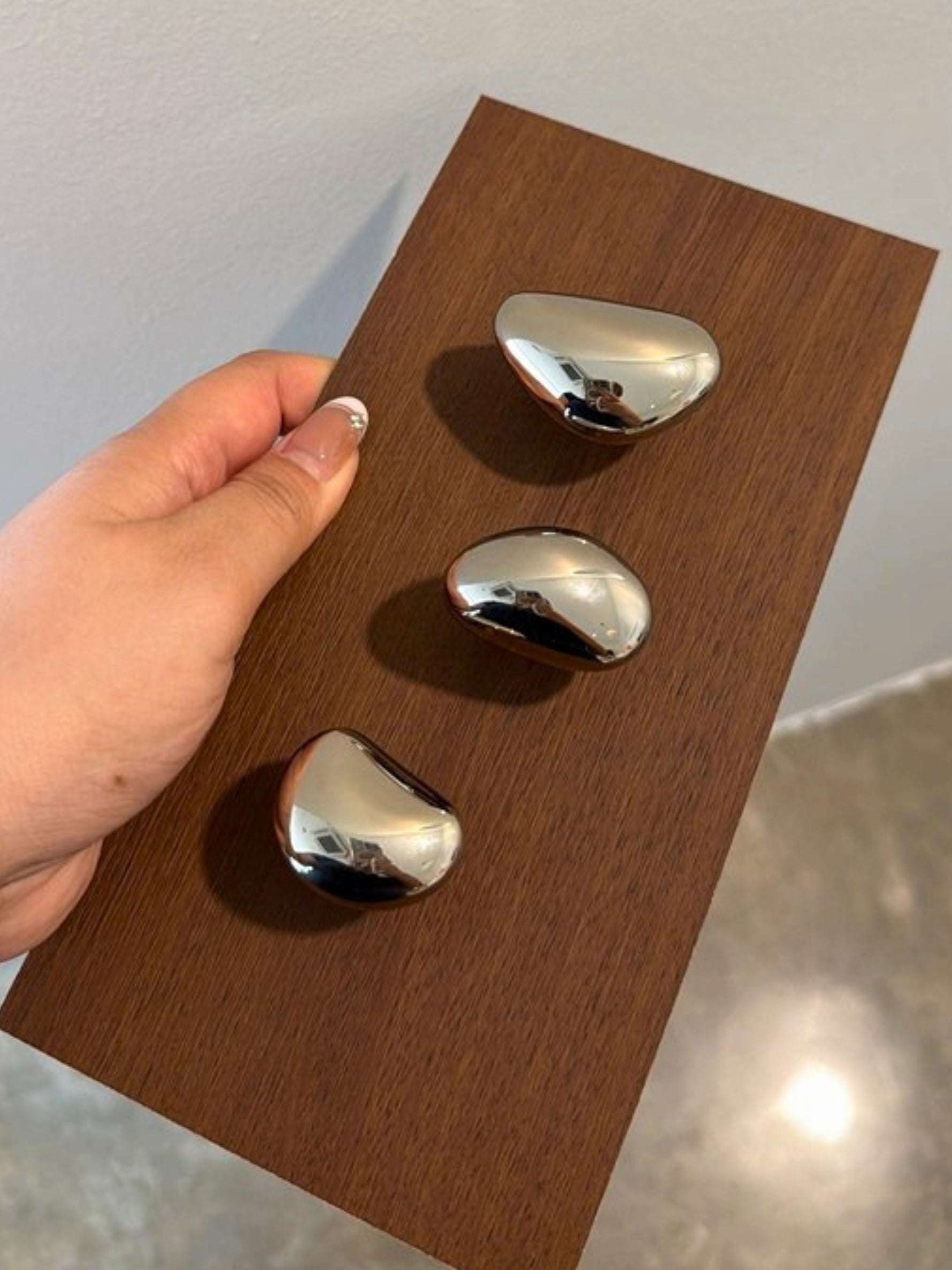 Hand holding three polished chrome pebble knobs of different sizes on wood panel
